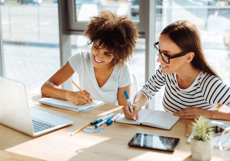 A couple of women sitting at a table writing in notebooks - Media Description