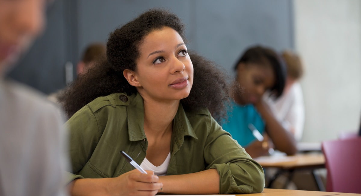 Woman sitting in a classroom at a desk, holding a pen and looking up.